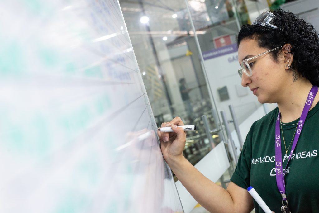 Colaboradora da Gali Energia escreve em quadro branco dentro da fábrica, utilizando caneta marcador. Ela usa óculos de proteção, crachá e uniforme com a frase “Movidos por Ideais”.