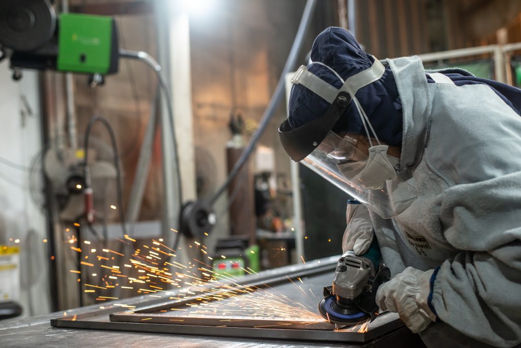 Colaborador do setor de solda realizando acabamento de peça metálica com esmerilhadeira, utilizando equipamentos de proteção individual como máscara facial, luvas e vestimenta de segurança, em ambiente de montagem e fabricação industrial.