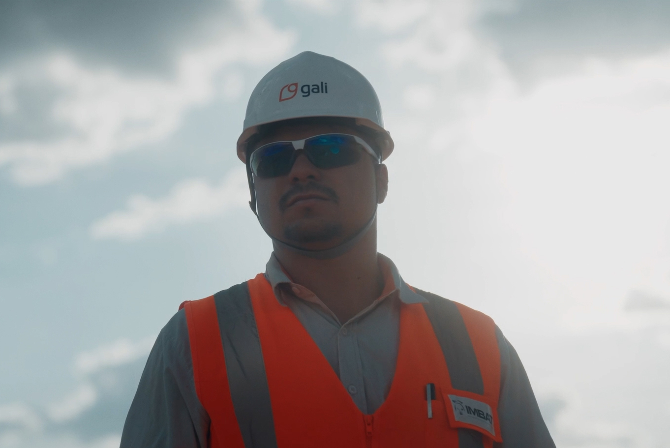 Profissional da Gali Energia com colete laranja e capacete branco, usando EPIs em campo sob o sol, representando atuação técnica segura no setor de energia.