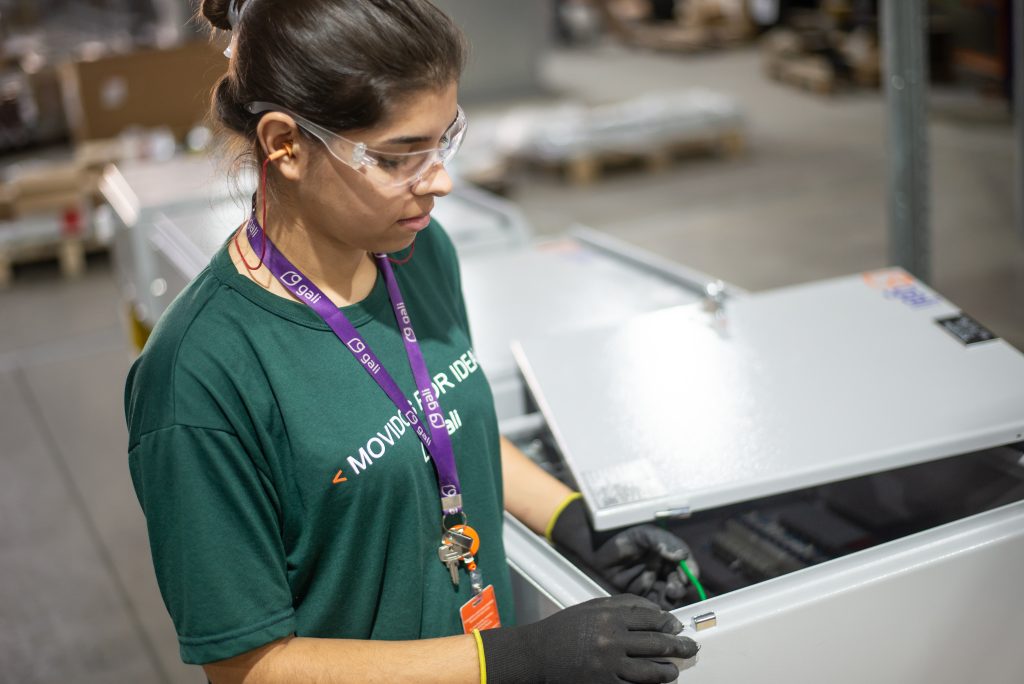 Colaboradora da Gali Energia manuseia painel elétrico em ambiente industrial, usando EPIs como óculos de proteção, luvas e crachá. A profissional está uniformizada e atua na montagem de equipamentos elétricos, representando a presença feminina na engenharia elétrica e no setor de energia sustentável.