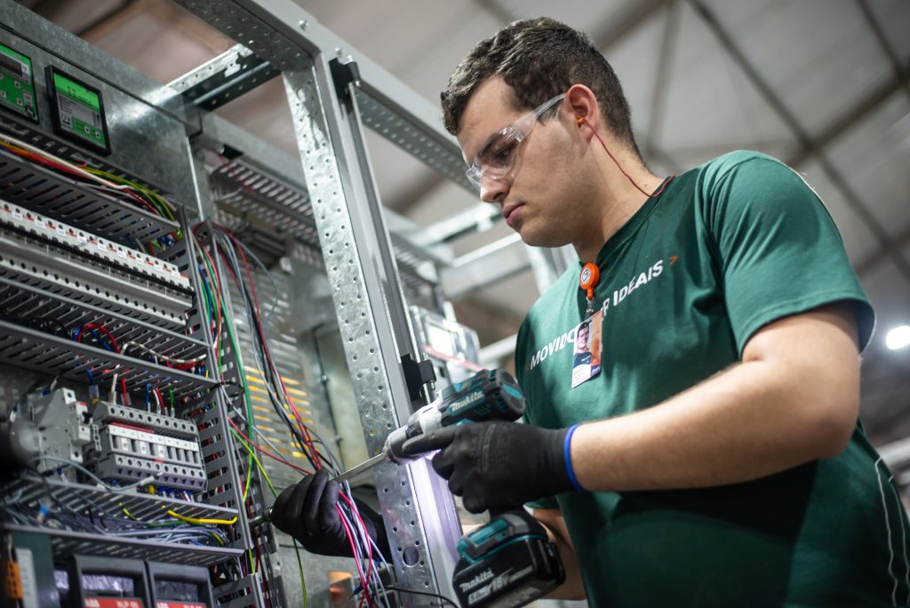 Colaborador da Gali Energia realiza montagem de painel elétrico industrial, utilizando parafusadeira e equipamentos de proteção individual, como luvas, óculos de segurança e protetor auricular. A cena representa o trabalho qualificado em instalações elétricas e automação, reforçando o compromisso da Gali com a excelência em engenharia e segurança no setor de energia sustentável.