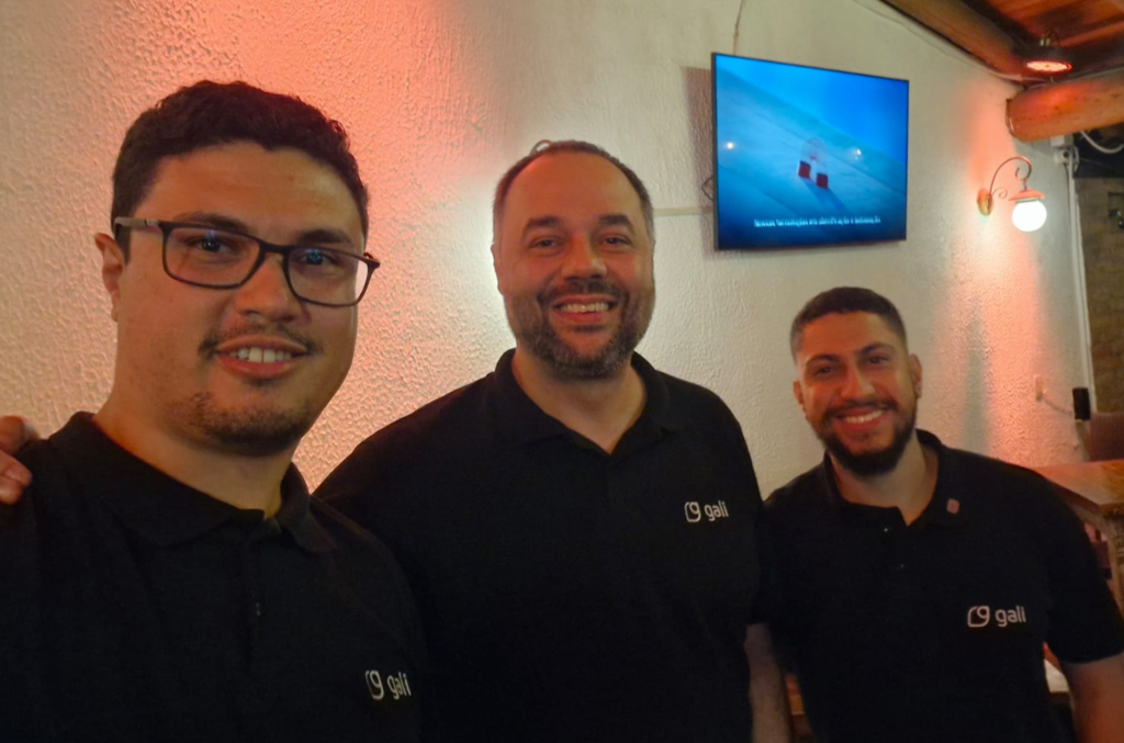 Três colaboradores da Gali Energia, todos sorrindo, posam juntos para uma foto em um ambiente interno com iluminação quente. Eles vestem camisetas pretas com o logo da empresa. Ao fundo, uma televisão exibe uma imagem azulada com texto, e a parede é decorada com luminárias. A imagem foi registrada durante o evento da ABB sobre tecnologias disruptivas na indústria.