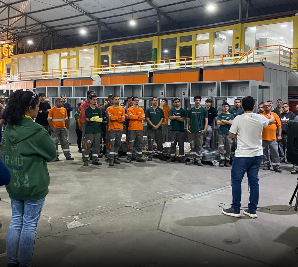 Colaboradores da Gali reunidos em área fabril durante encontro do Projeto Galileu. O diretor operacional, Gabriel Daré, aparece de costas enquanto apresenta os resultados do semestre e o planejamento estratégico de 2025. Ao fundo, colunas de um cubículo de média tensão e o mezanino administrativo da empresa.