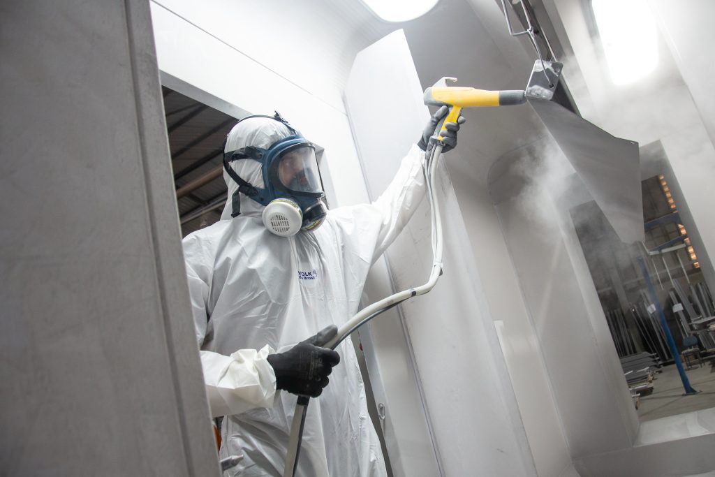 Profissional da Gali Energia realiza aplicação de pintura eletrostática em peça metálica dentro da cabine de pintura, utilizando equipamento de alta precisão. Vestindo macacão de proteção e máscara com filtro, o colaborador garante segurança, qualidade e acabamento uniforme no processo industrial da fábrica em Bauru (SP).