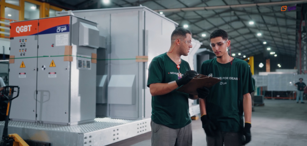 Dois colaboradores da Gali Energia, usando equipamentos de proteção individual (EPI) como luvas e óculos de segurança, estão em uma fábrica analisando uma prancheta com anotações. Ao fundo, é possível ver um painel elétrico QGBT e parte de um eletrocentro em montagem, destacando o ambiente industrial e a produção de painéis de baixa tensão.