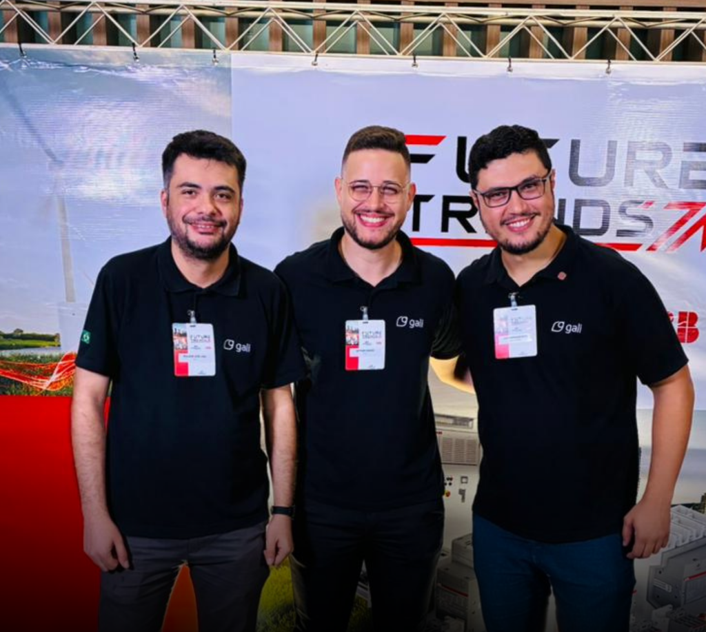 Três integrantes da equipe comercial da Gali posam sorridentes durante o evento Future Trends, promovido pela Dimensional Brasil e ABB Brasil, em Bauru (SP). Eles vestem camisetas pretas com o logotipo da Gali e crachás de identificação, em frente a um painel com o nome do evento.