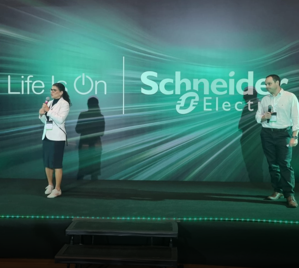 Duas pessoas em um palco durante apresentação do evento Innovation Talk, promovido pela Schneider Electric. Ao fundo, há um grande painel verde com o logotipo da Schneider Electric e a frase “Life is On”. A iluminação é suave e o cenário tem efeito de movimento em tons de verde e preto.