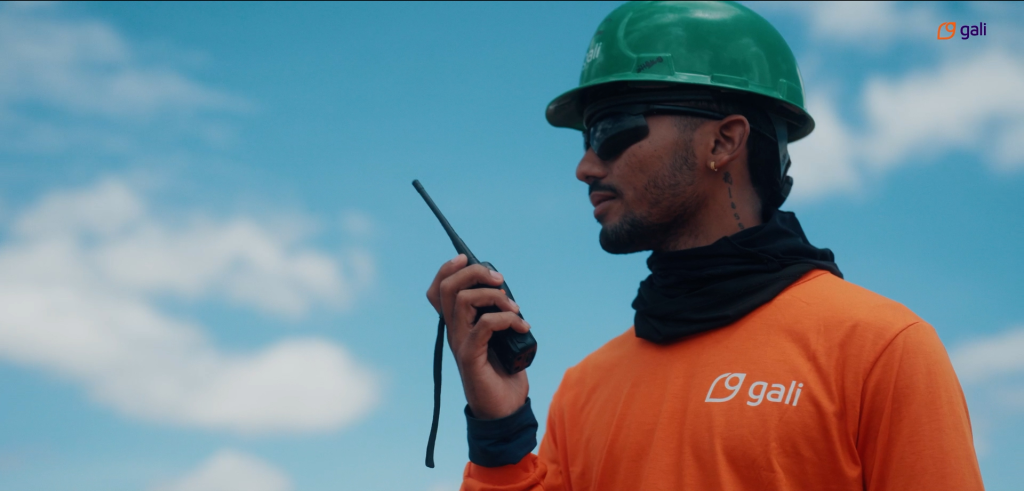 Profissional da Gali Energia, usando capacete verde e uniforme laranja, se comunica por rádio em uma obra a céu aberto.