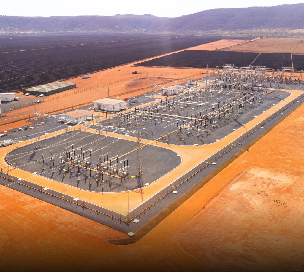 Vista aérea da Subestação Elevadora Várzea da Palma 4, localizada em Minas Gerais. A imagem mostra a estrutura completa da subestação, com seus equipamentos de alta tensão, pátios energizados e área cercada por alambrado. Ao fundo, é possível ver o Complexo Fotovoltaico Hélio Valgas, com centenas de painéis solares distribuídos pelo terreno.