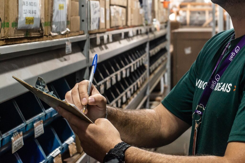 Colaborador da Gali está conferindo as prateleiras do almoxarifado, segurando em mãos uma prancheta e escrevendo nela com uma caneta. O colaborador utiliza uniforme e crachá da Gali e um relógio na mão esquerda.