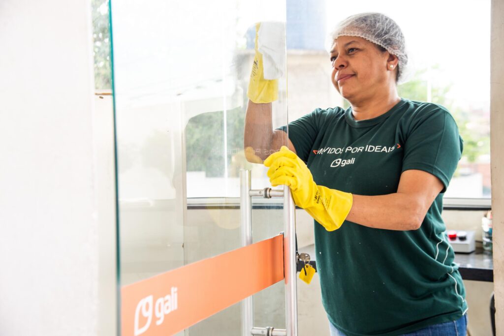 Profissional de serviços gerais da Gali limpando porta de vidro com luvas amarelas.