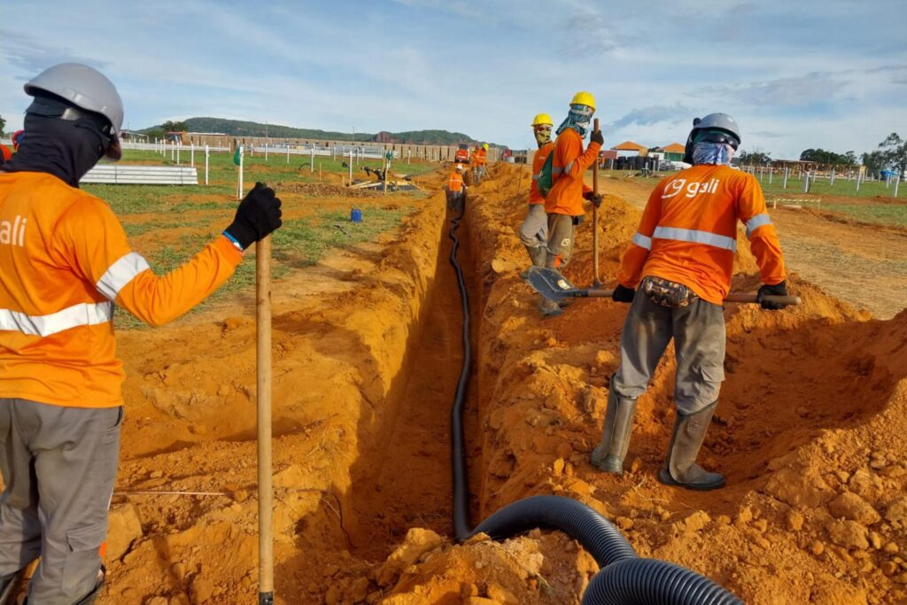 Equipe da Gali trabalhando em uma obra de energia solar, escavando valas e instalando tubulações elétricas em um campo aberto, utilizando EPIs completos e uniformes de segurança.