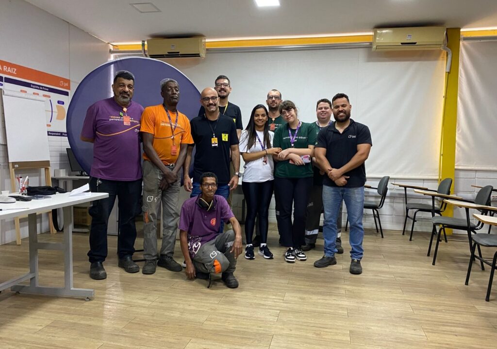 Equipe posando em sala de aula corporativa, com participantes de diferentes áreas lado a lado. Espaço interno com móveis simples e clima de colaboração.