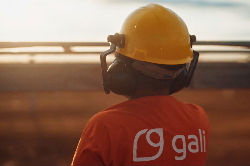 Colaborador da Gali usando capacete amarelo, óculos e protetor auricular durante atividade em obra de energia, com cenário ao ar livre ao pôr do sol.