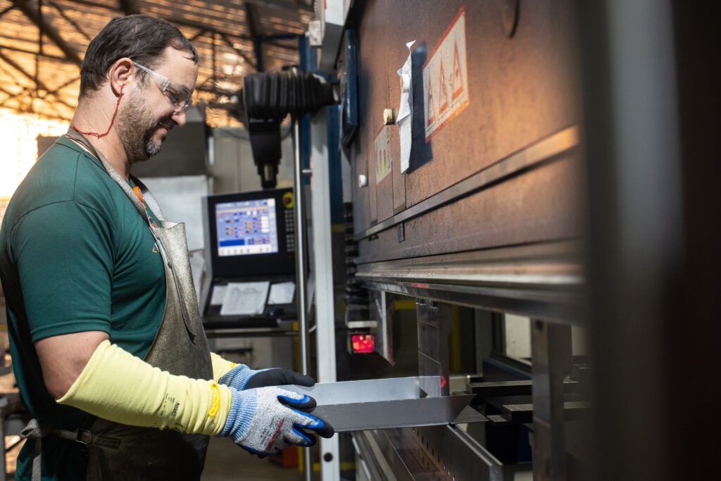 Profissional da Gali opera máquina de dobra de chapas metálicas em ambiente industrial, utilizando EPIs e seguindo padrões de segurança.