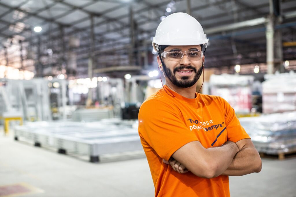 Colaborador da Gali em ambiente industrial, usando capacete e óculos de proteção, posa com braços cruzados em fábrica de estruturas metálicas.