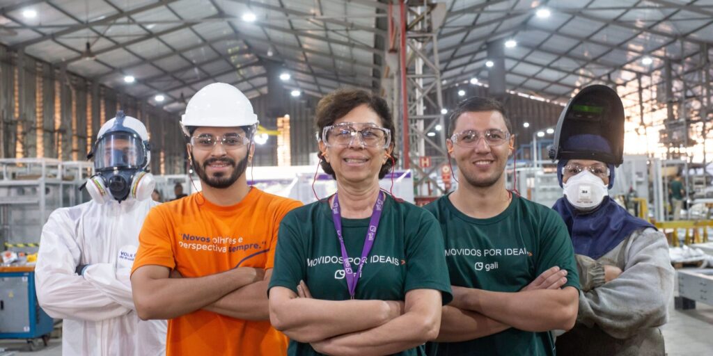 Equipe industrial diversa da Gali Energia posa em fábrica com equipamentos de proteção individual (EPIs), incluindo capacetes, óculos, máscaras e uniforme técnico.