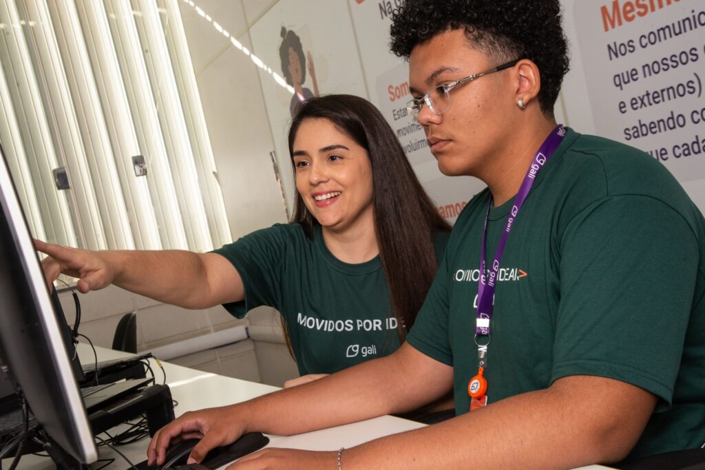Dois colaboradores da Gali em ambiente corporativo trabalham juntos em frente ao computador, com uma profissional apontando para a tela enquanto orienta o colega.